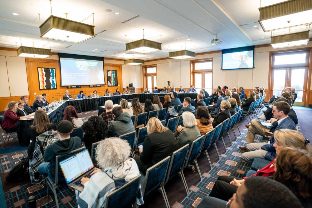 A U‑shaped panel of about a dozen people seated at a long table conducts a formal meeting or board session in a conference room, with two projection screens showing slides at the front. Dozens of attendees sit in rows facing the panel.